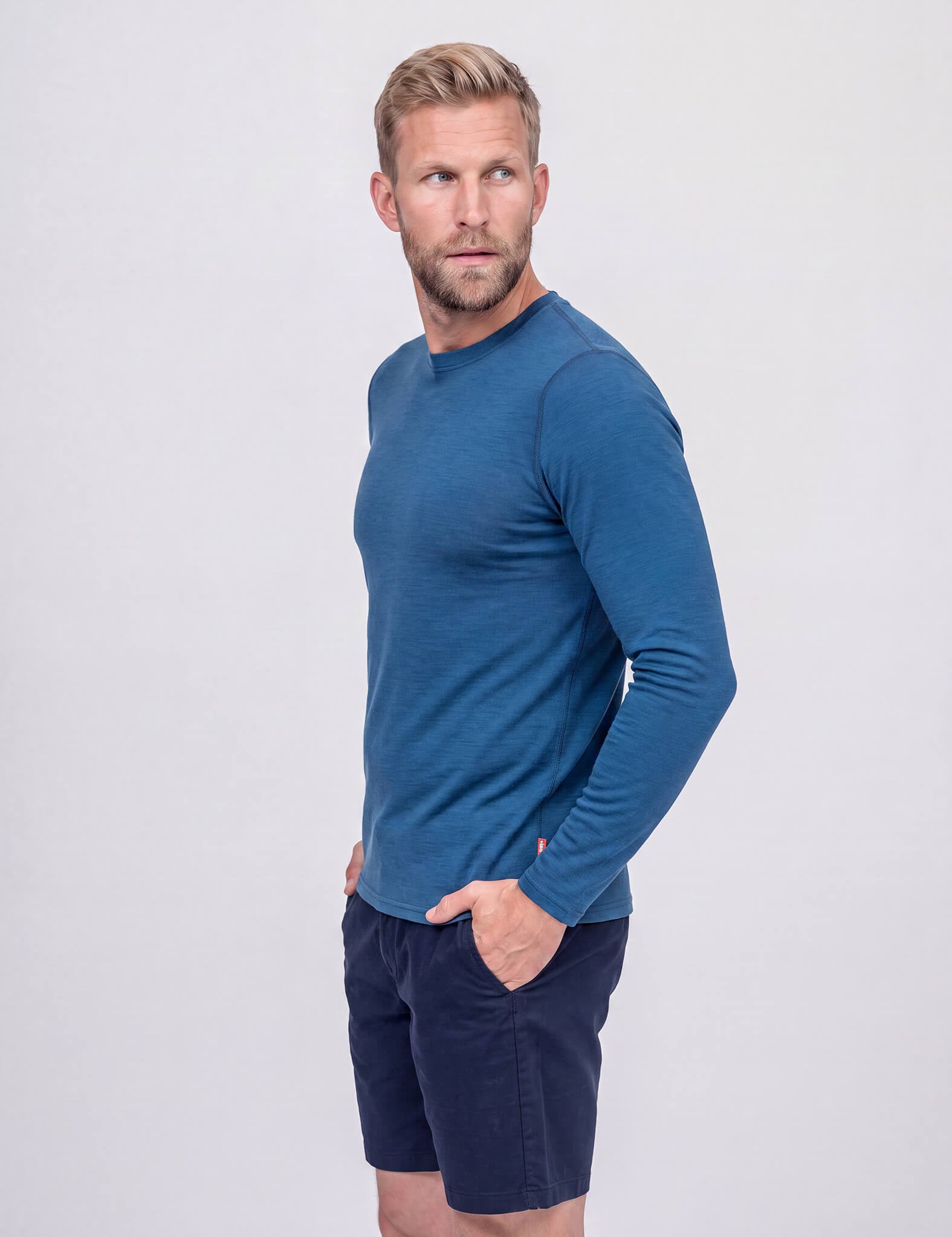 A man with blond hair and a beard wears the Crew Neck – Classic Merino 160 Men in Ensign Blue and dark shorts. He stands with his hands in his pockets, looking to his left, against a plain light background.
