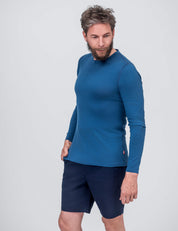 A man with short brown hair and a beard wears the Crew Neck – Classic Merino 160 Men in Ensign Blue and navy shorts, standing against a plain light gray background, looking down to his left with one hand in his pocket.