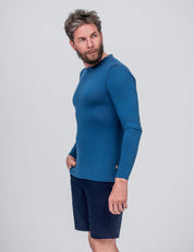 A man with short, gray hair and a beard wears the Crew Neck – Classic Merino 160 Men long-sleeve blue shirt and dark shorts, standing against a light gray background with one hand in his pocket—perfect for daily wear.