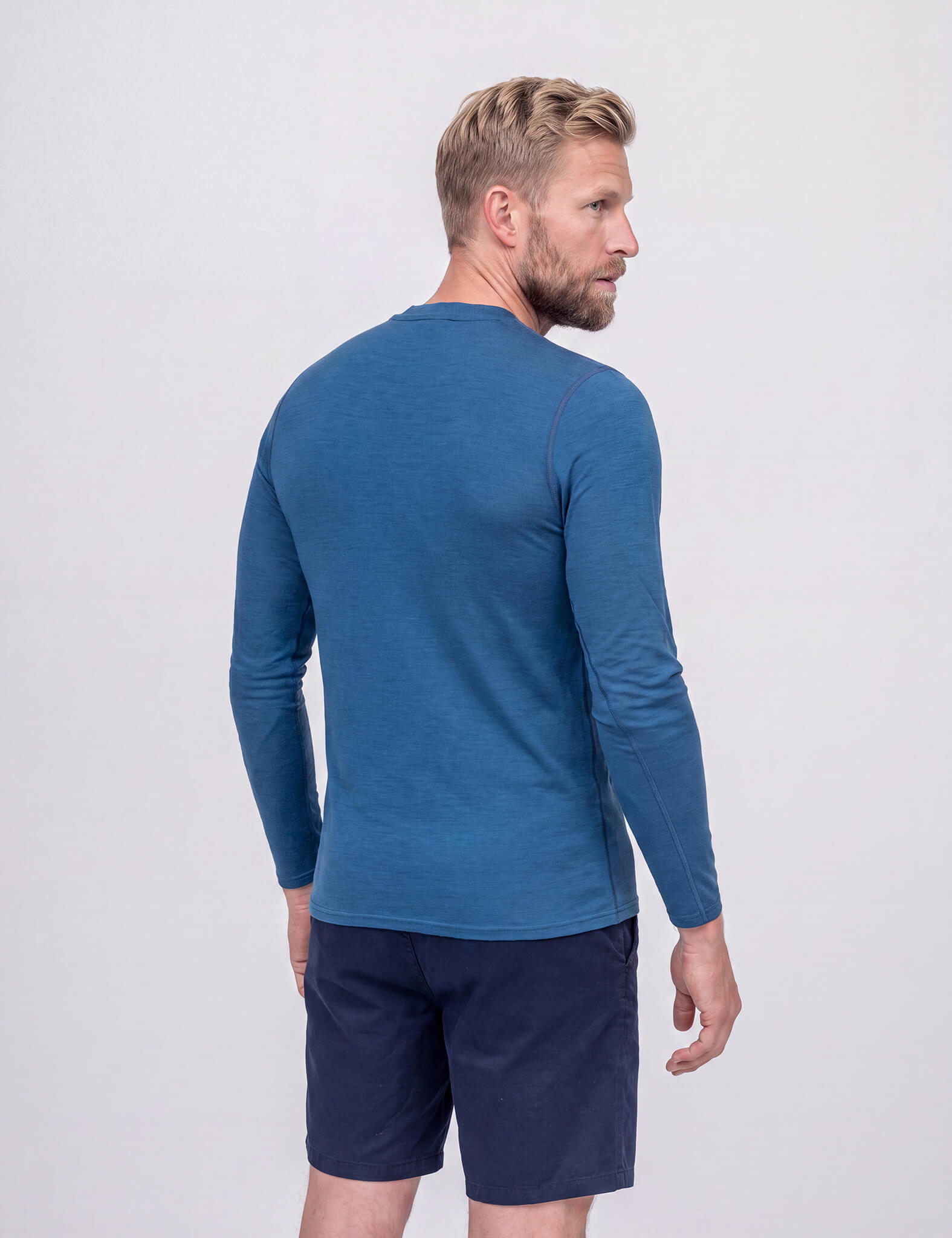 A man with short blond hair is standing and facing away, wearing the Crew Neck – Classic Merino 160 Men in blue and navy shorts against a plain light background.