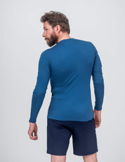 A man with brown hair and a beard wears the Crew Neck – Classic Merino 160 Men long-sleeve in blue and navy shorts, standing slightly turned away against a plain light gray background—ideal for everyday use.