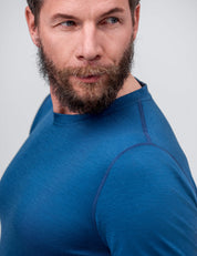 A light-skinned, bearded man wears the Crew Neck – Classic Merino 160 Men in blue, gazing thoughtfully to his left against a neutral background—a stylish, fitted long-sleeve layer ideal for everyday wear.