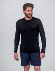A man with short brown hair and a beard is wearing the Crew Neck – Classic Merino 160 Men in black, paired with navy blue shorts, and stands against a plain light gray background—ideal for everyday wear.
