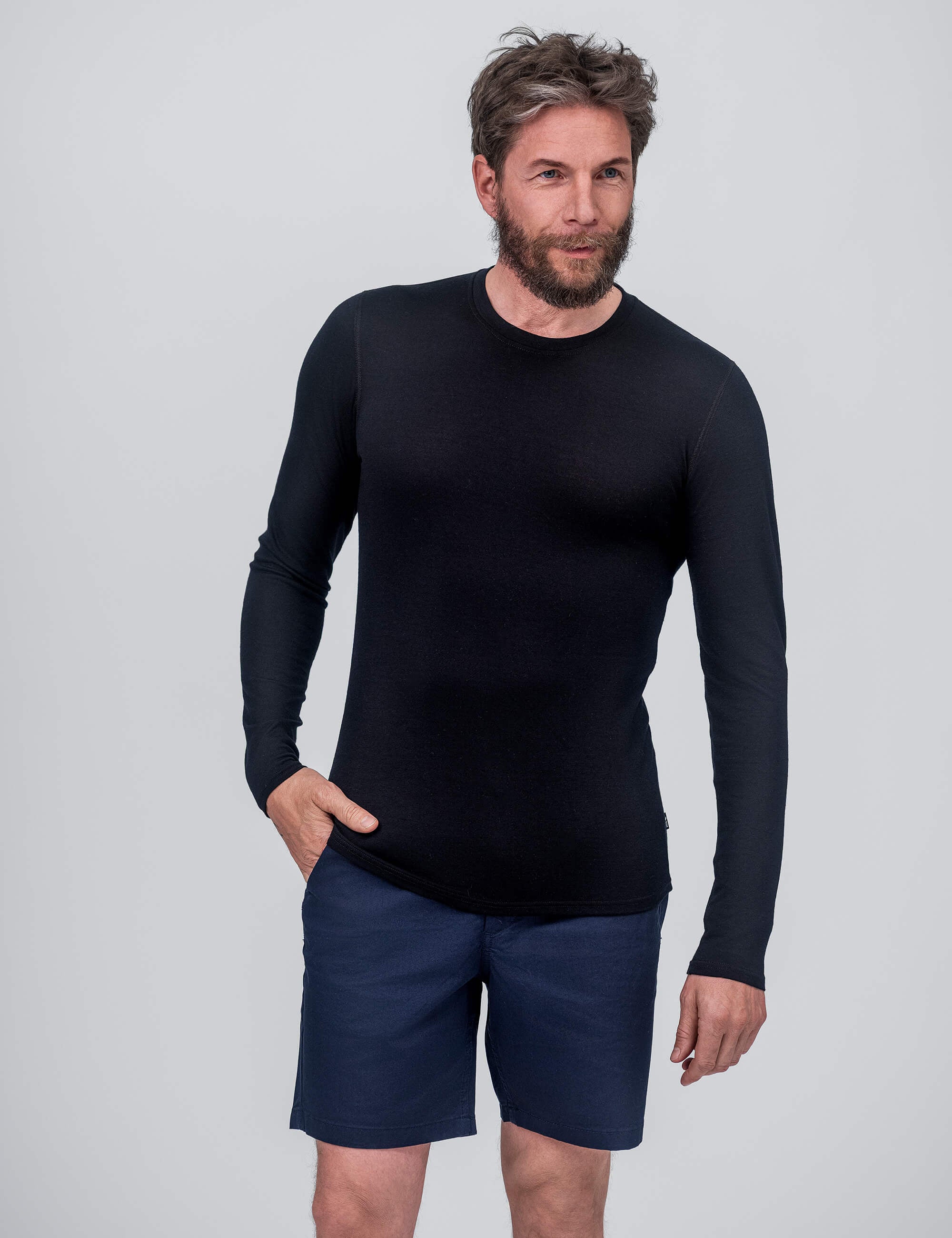 A man with a beard and tousled hair wears the Crew Neck – Classic Merino 160 Men in black and navy blue shorts, standing against a plain light gray background, one hand in his pocket, smiling slightly.