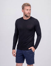A man with blond hair and a beard wears the Crew Neck – Classic Merino 160 Men in black, paired with navy blue shorts, standing against a plain light background, looking to his left with one hand in his pocket—perfect for everyday use.