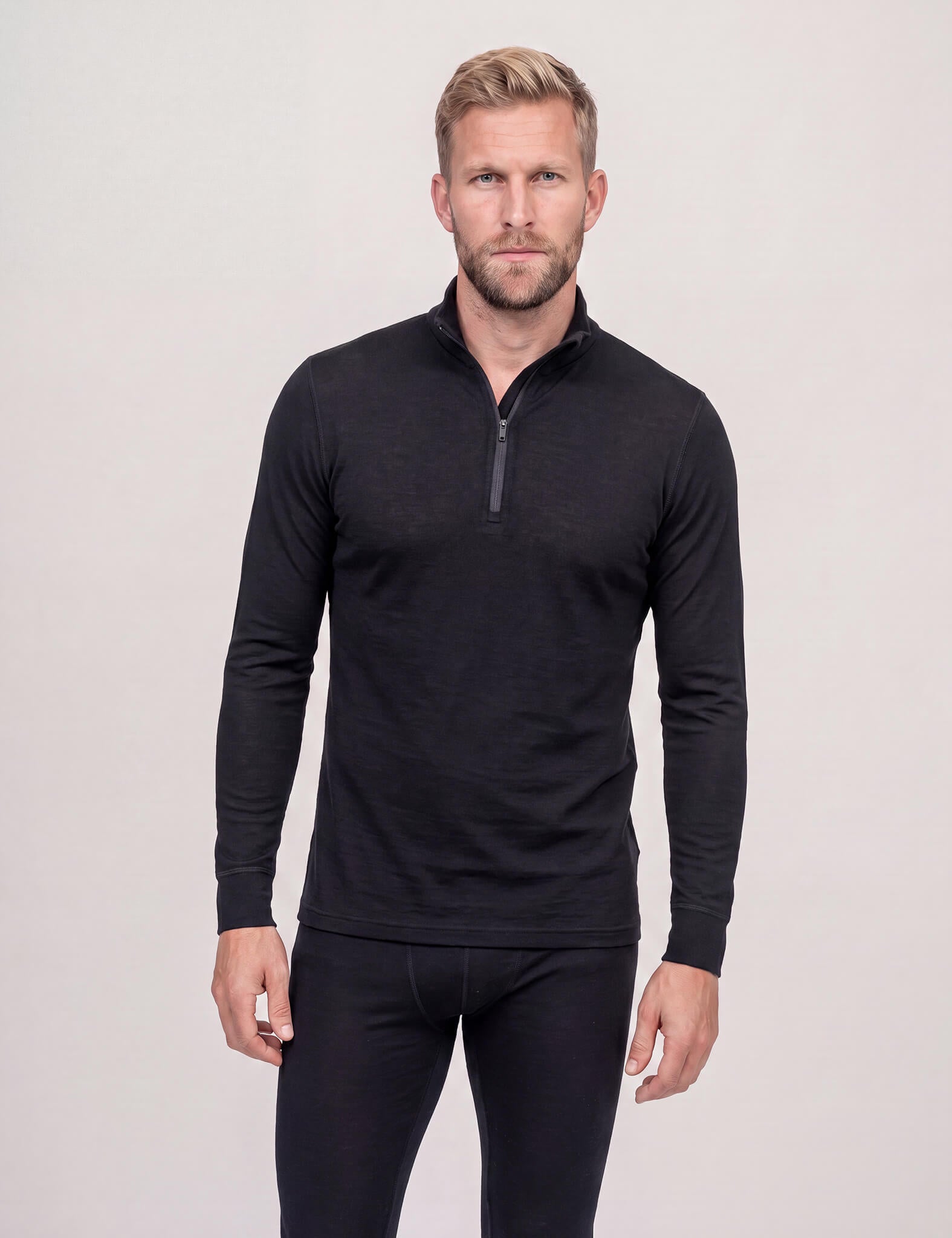 A man with blond hair and a beard is wearing the Half Zip – Classic Merino 160 Men in black, along with matching black pants, standing against a plain light background. This activewear provides natural temperature regulation for all-day comfort.