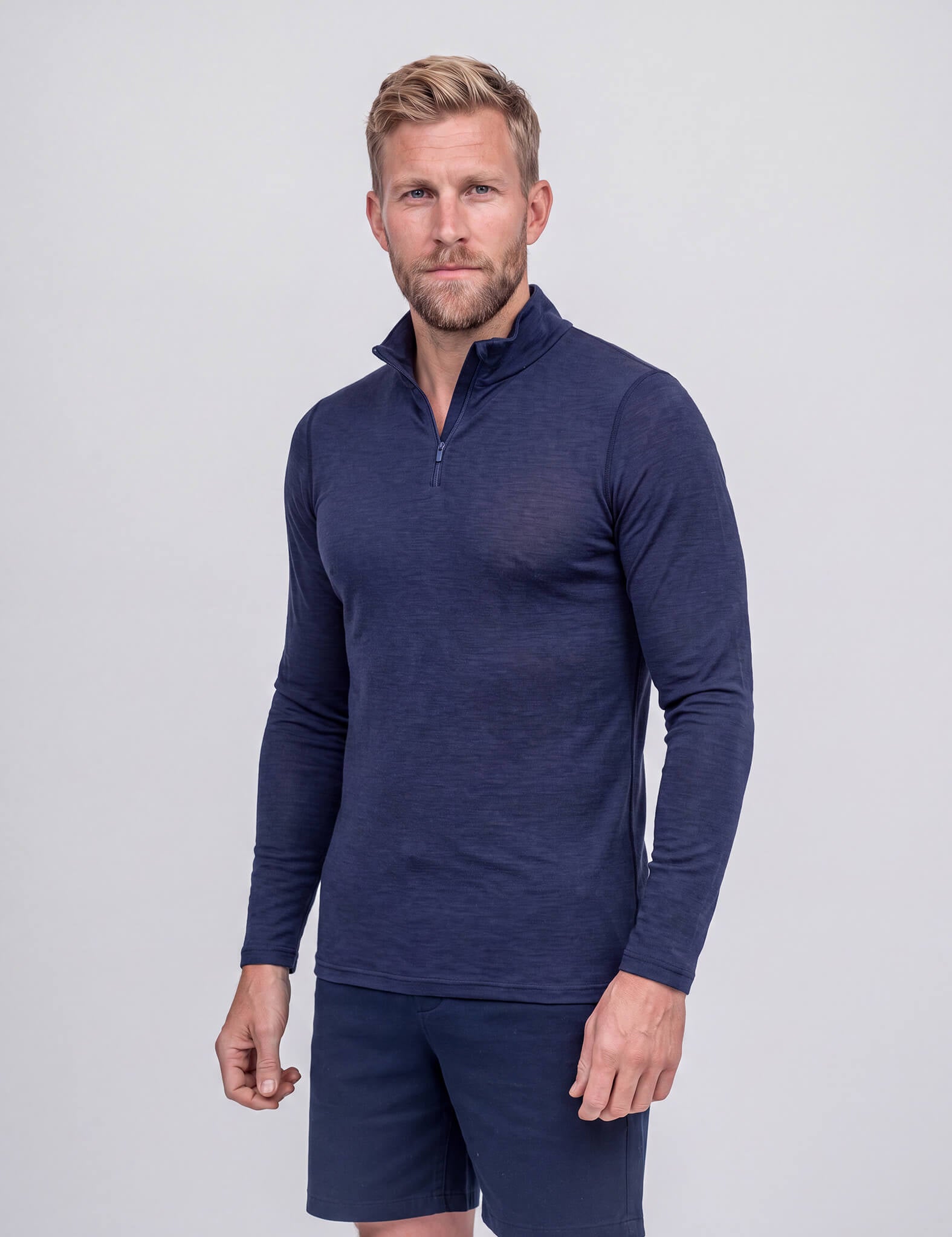 A man with short blond hair and a beard wears the Half Zip – Classic Merino 160 Men in navy, along with matching activewear shorts, standing against a plain light gray background.