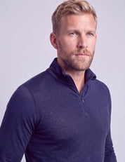 A man with blond hair and a beard, dressed in the Half Zip – Classic Merino 160 Men in dark blue, stands against a light purple background, gazing slightly to his left with a neutral expression.