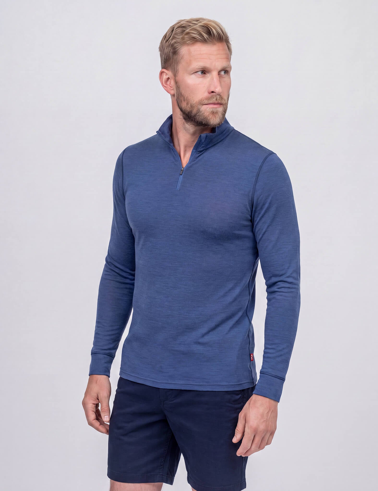 A man stands against a plain, light background, looking off to his right with one hand at his side while wearing the Half Zip – Classic Merino 160 Men in Ensign Blue and dark blue shorts. This activewear piece features natural temperature regulation for all-day comfort.