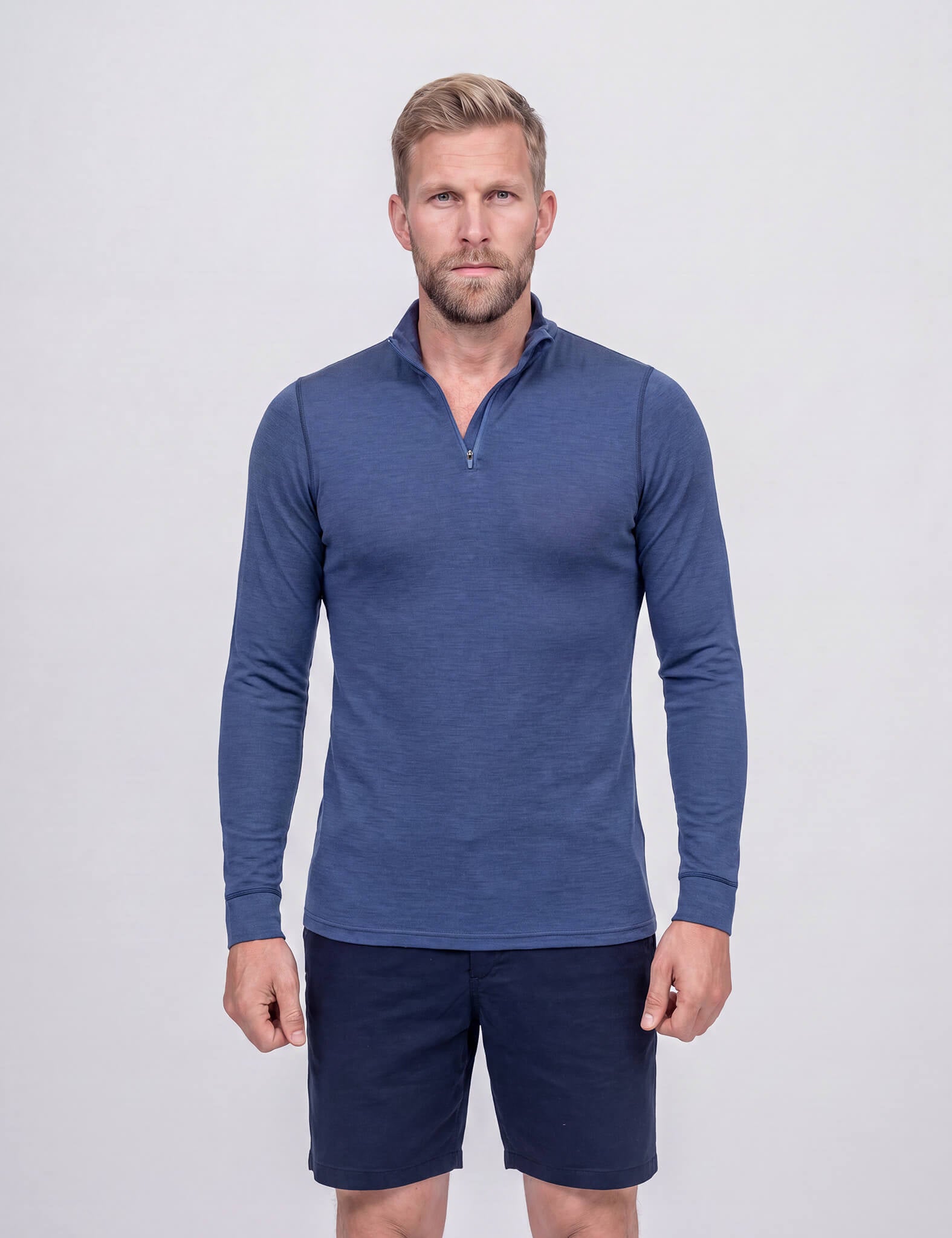 A man wearing the Half Zip – Classic Merino 160 Men in blue and navy shorts stands in front of a plain light-gray background, facing forward with a neutral expression.