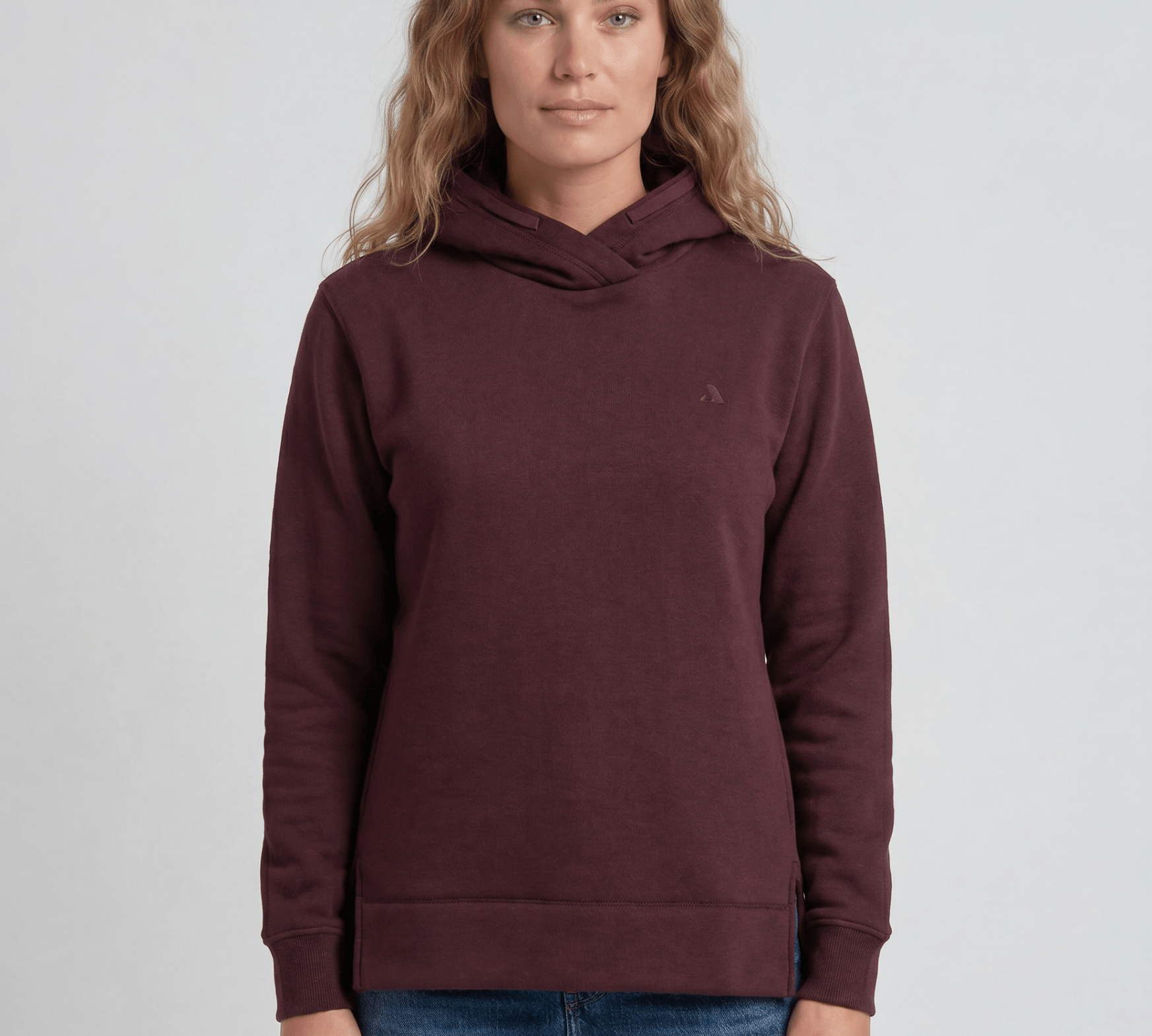 A woman with wavy, light brown hair wears the maroon Hoodie Merino – AirWool 280 Women and blue jeans, standing against a plain light gray background and facing the camera—ideal for cold weather.