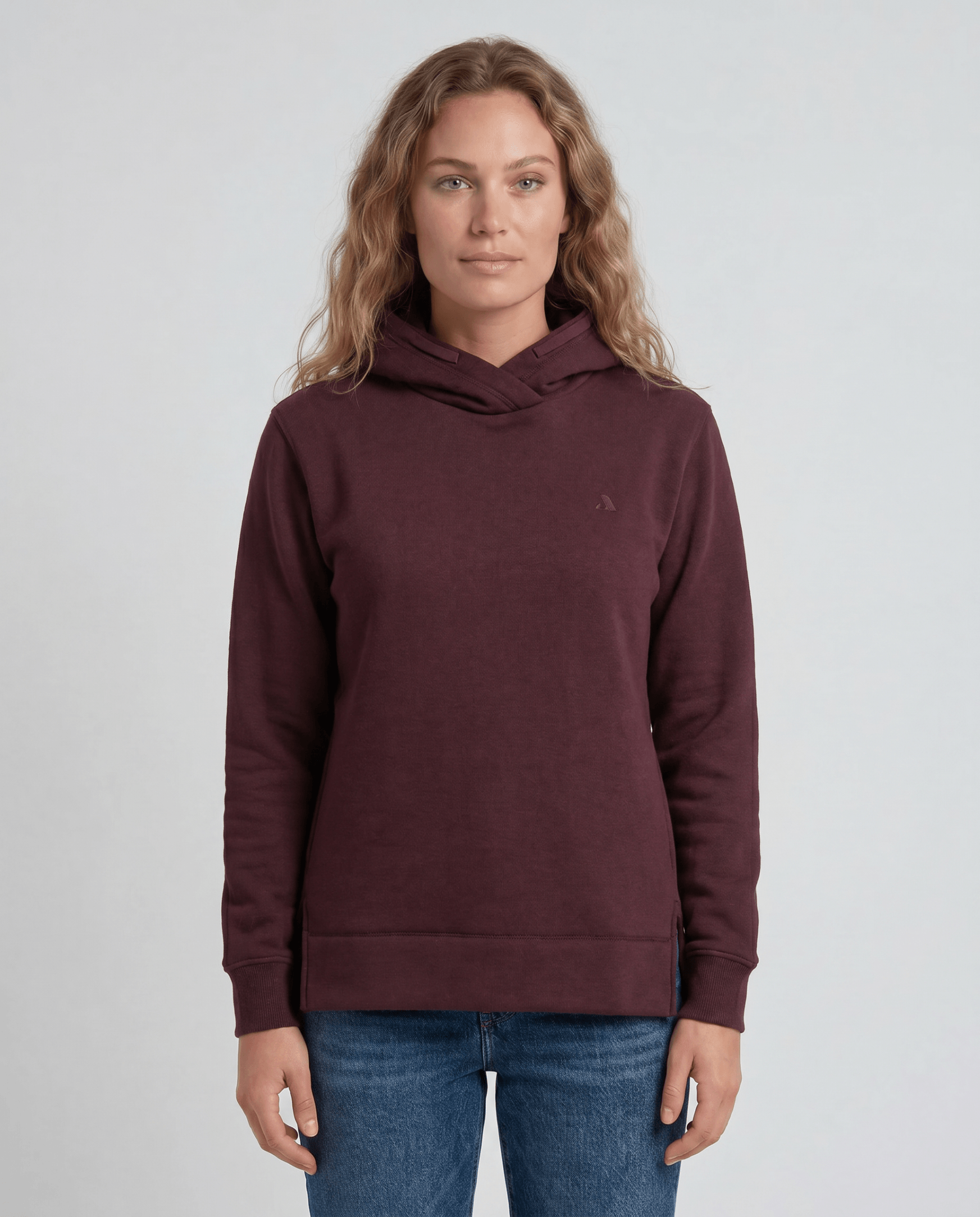 A woman with wavy, light brown hair wears the maroon Hoodie Merino – AirWool 280 Women and blue jeans, standing against a plain light gray background and facing the camera—ideal for cold weather.