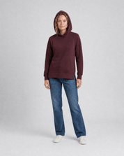 A woman wears the Hoodie Merino – AirWool 280 Women with the hood up, paired with blue jeans and white sneakers against a light gray background—showcasing this stylish, insulating hoodie ideal for cold weather.