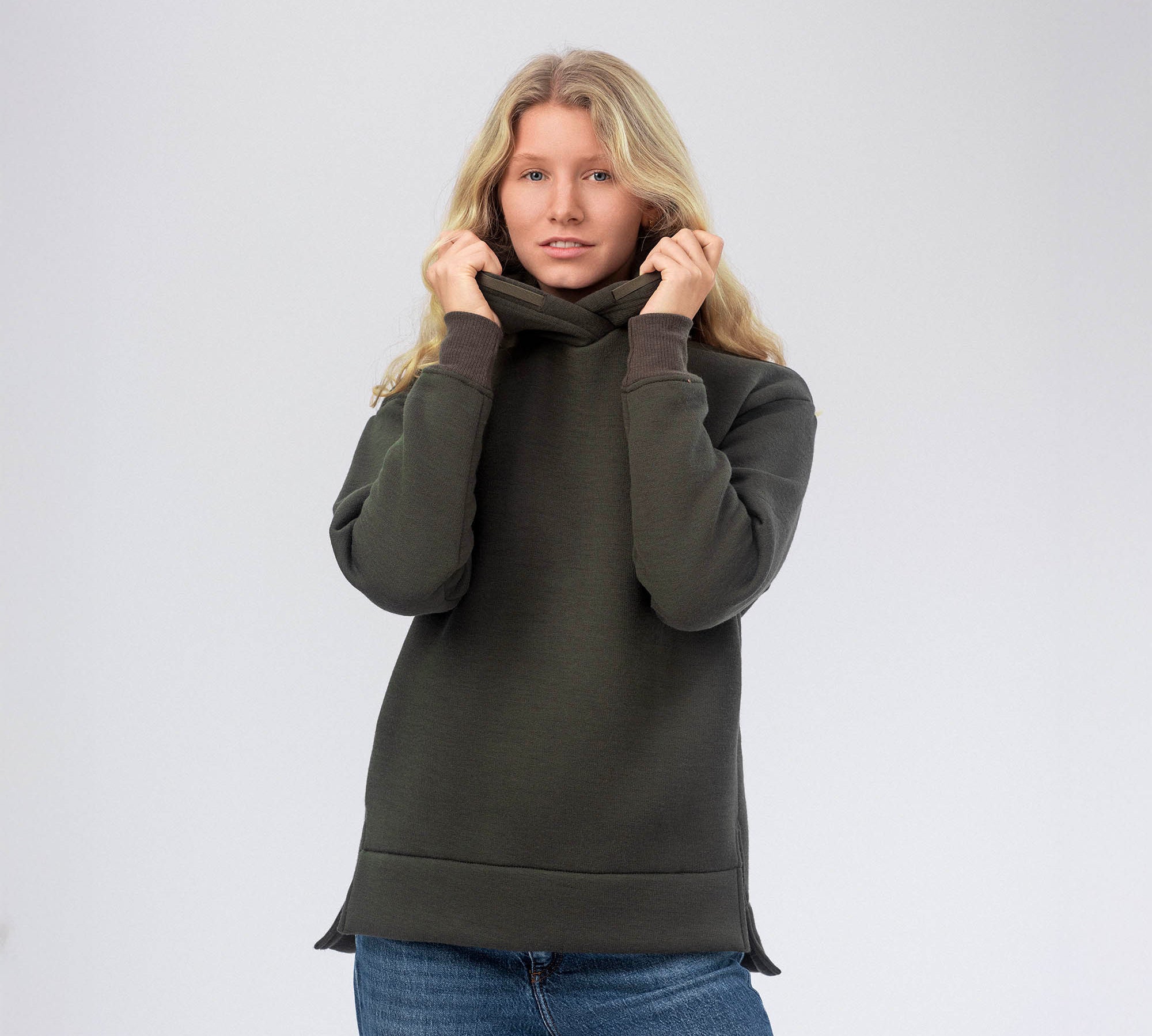 A woman with long blonde hair wears the Forest Night Hoodie Merino – AirWool 280 Women, holding its collar with both hands while standing against a plain light background—ideal for cool to cold conditions.