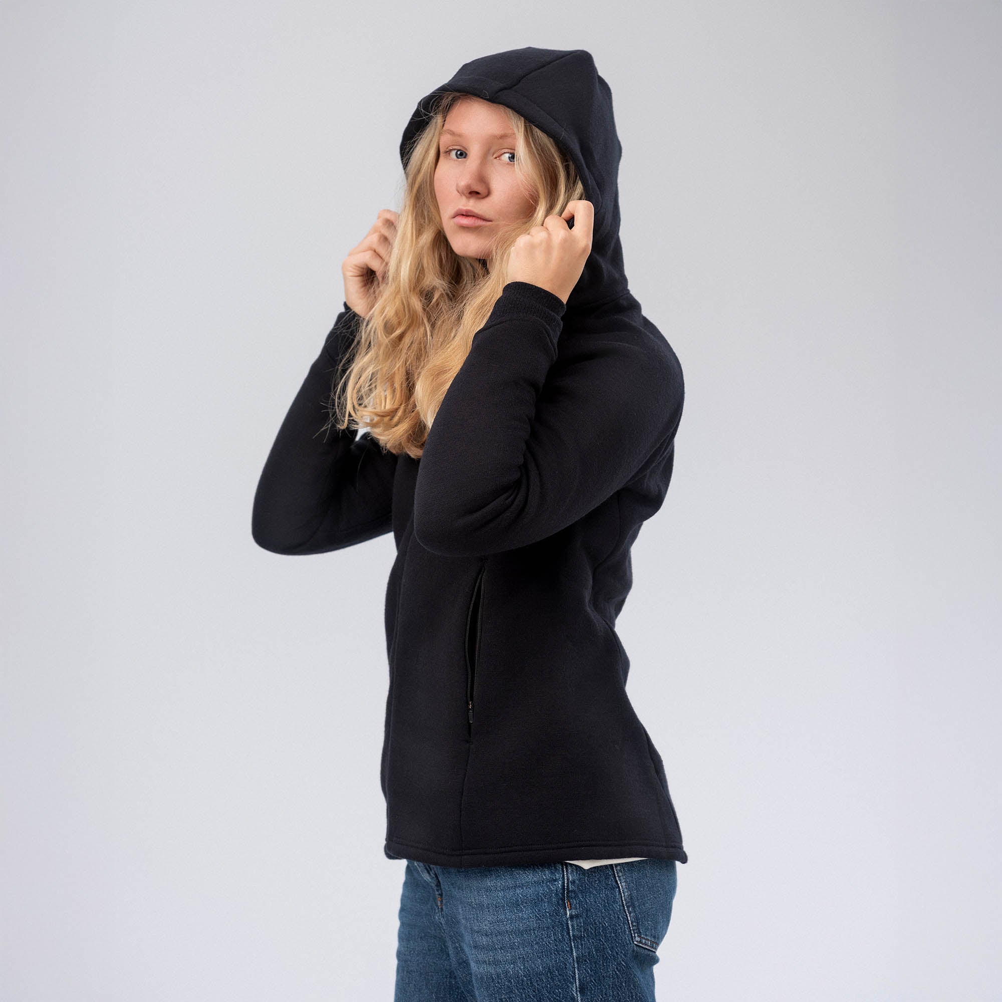 A woman with long blonde hair wears the Hooded Jacket Merino – AirWool 280 Women in black and blue jeans, standing sideways, holding the jacket with both hands, looking ahead with a neutral expression against a plain light background.