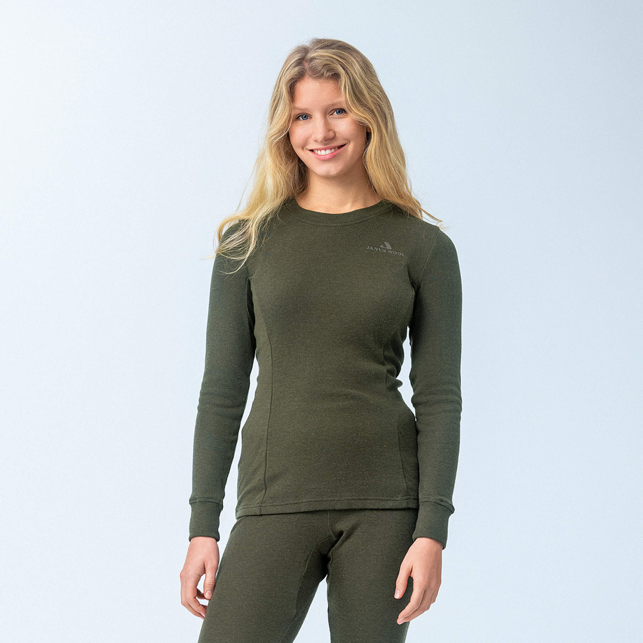 A woman with long blonde hair smiles while wearing the Jersey – Classic Merino 215 Women set in Forest Night, a breathable and warm merino wool long-sleeve top and matching pants, against a plain light background.