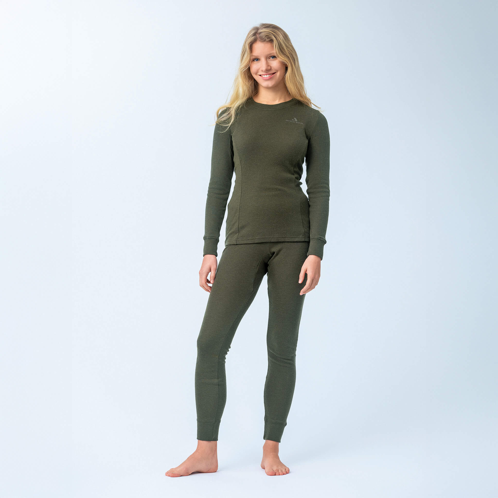 A woman with long blonde hair stands barefoot, smiling, and wearing the Jersey – Classic Merino 215 Women set—breathable dark green merino wool top and leggings—against a plain light background.