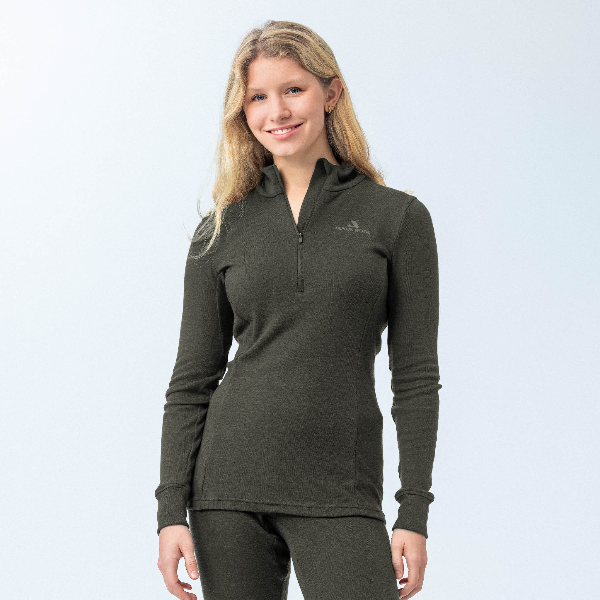 A woman with long blonde hair smiles in the Polo Zip – Classic Merino 215 Women, a fitted dark green merino wool polo and matching pants, standing against a light background that highlights the outfit’s warmth and breathability.