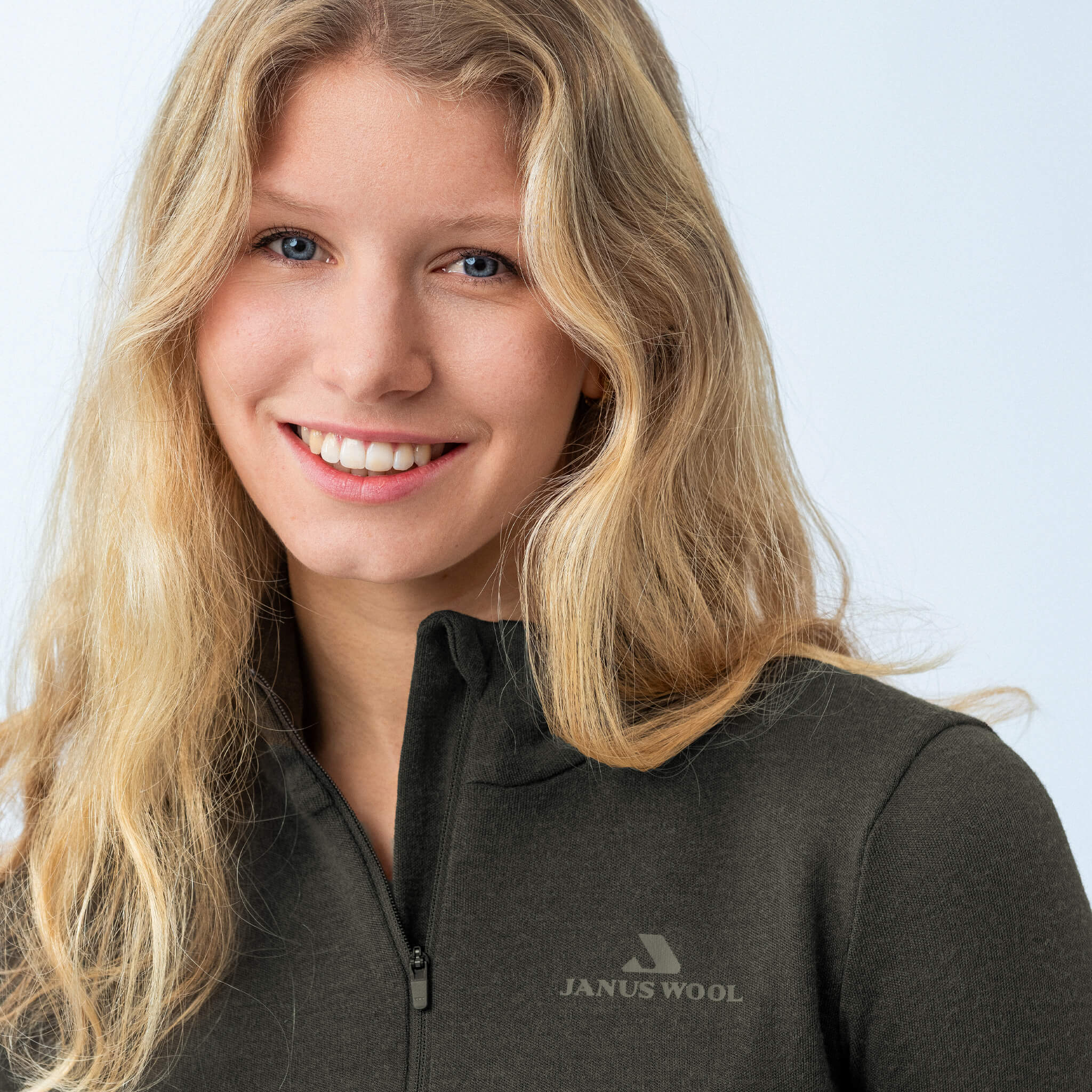 Smiling woman with long blonde hair wears the Polo Zip – Classic Merino 215 Women, featuring a logo, against a light background—showcasing its warmth and breathability.