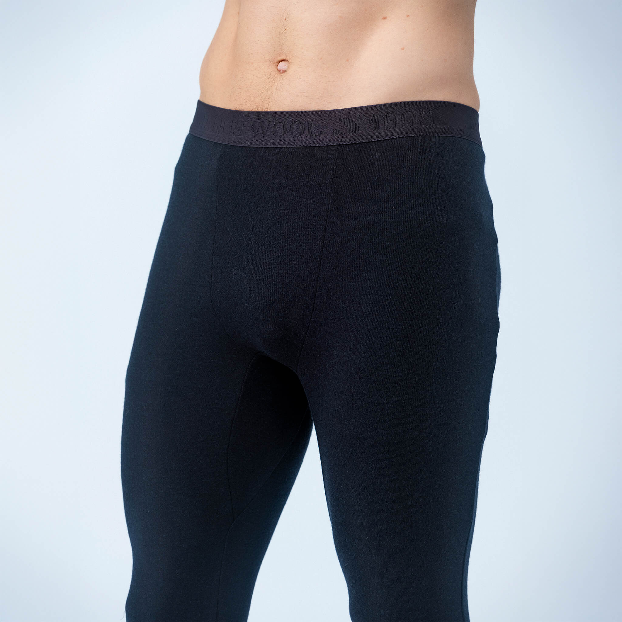 A man wears Classic Merino 215 Men leggings in black, shown waist to mid-thigh against a light blue background. The visible waistband displays text, emphasizing the leggings’ warmth and breathability.