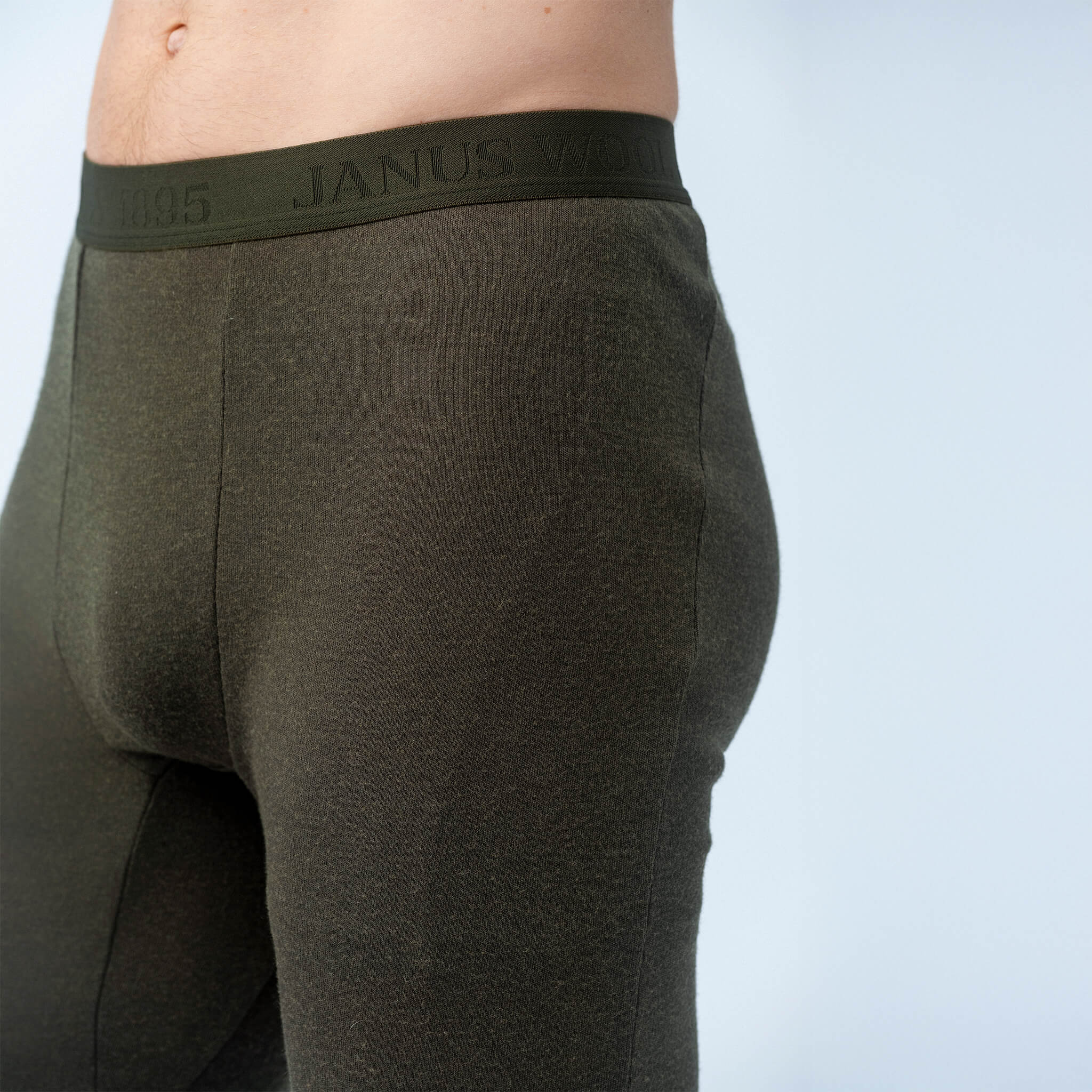 A close-up shows a person wearing Leggings – Classic Merino 215 Men in dark green with an elastic waistband, highlighting their warmth and breathability against a light background. The shot focuses on the lower torso and upper legs.