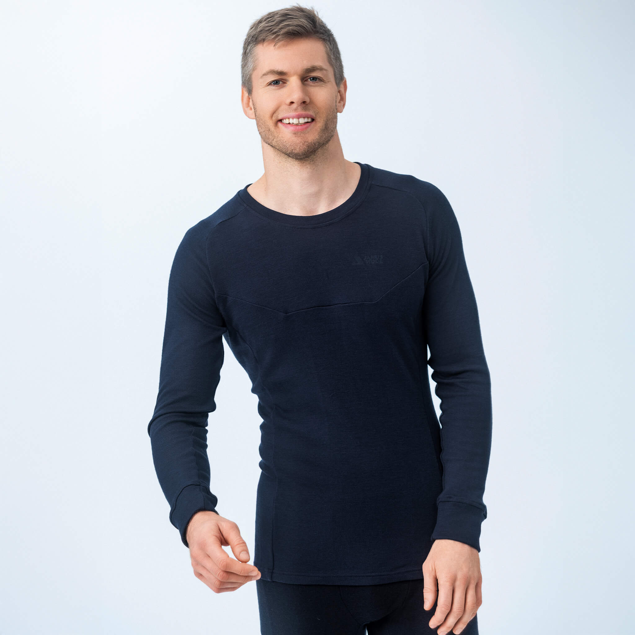 A man with short light brown hair smiles while wearing the Jersey – Classic Merino 215 Men in black, paired with black pants, standing against a plain, light background.