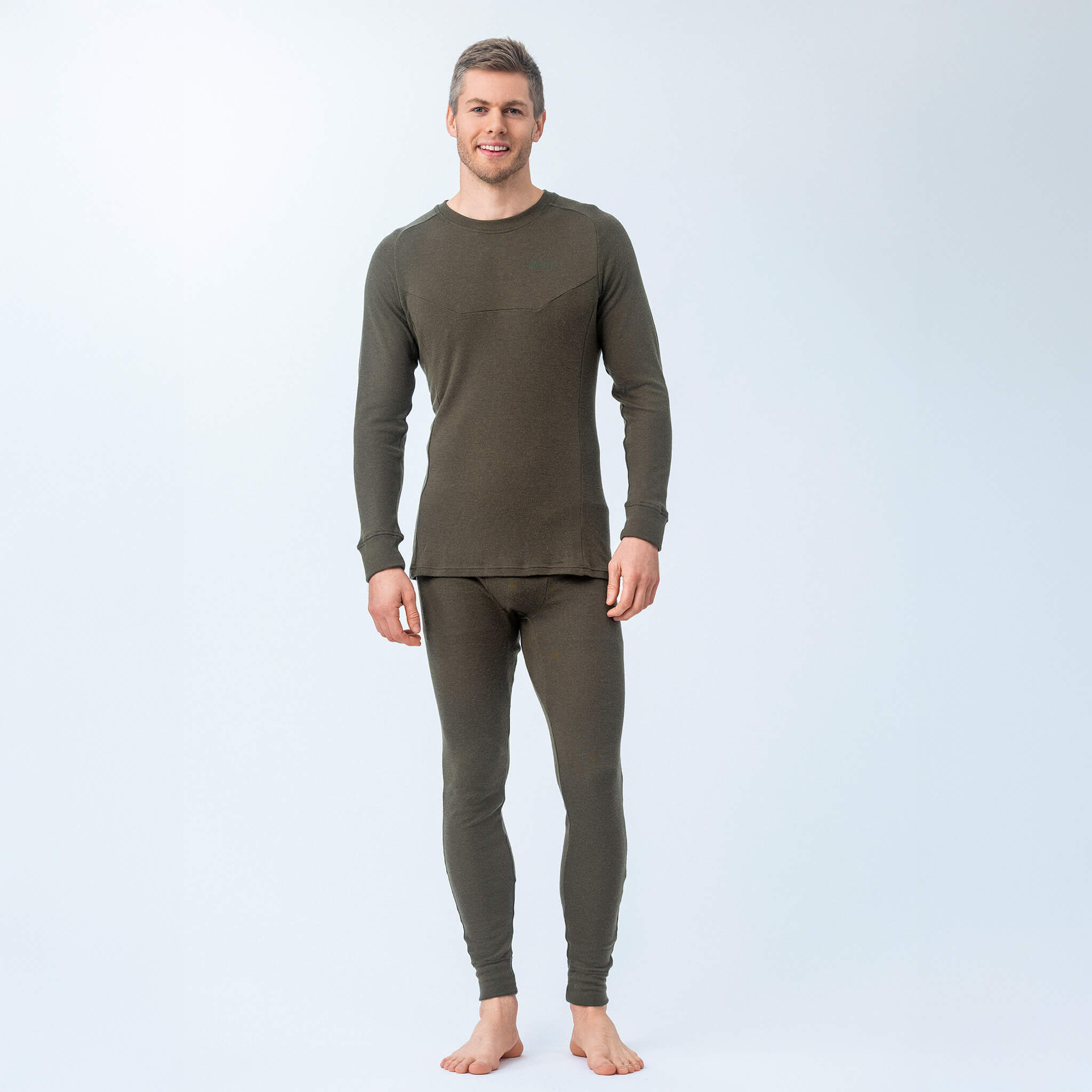 A man stands barefoot against a light background, facing forward and smiling in the Jersey – Classic Merino 215 Men, an olive green matching set with long sleeves and pants made from soft jersey material.
