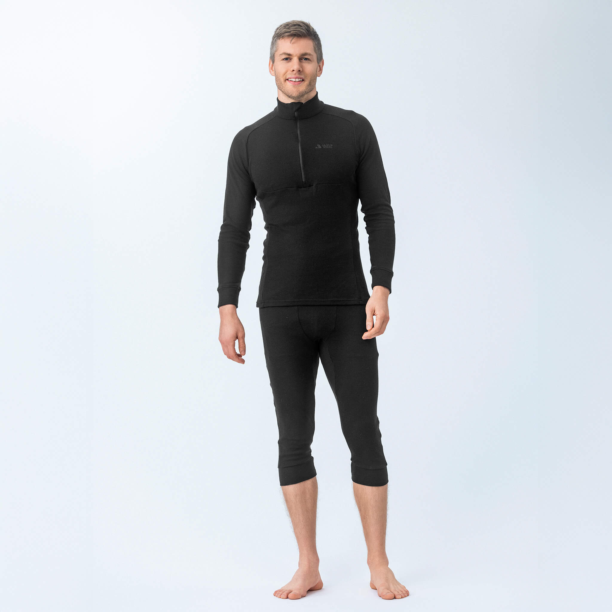 A man stands barefoot against a plain light background, smiling and facing forward, wearing the Polo Zip – Classic Merino 215 Men long-sleeve black top and matching three-quarter length bottoms.