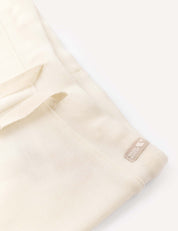 Soft cream-colored AirWool pants featuring a visible textured finish, drawstring loops, and a small sewn label displaying “100% ORGANIC COTTON” alongside a heart icon. These relaxed-fit Pants – Merino Baby AirWool are styled against a light, neutral background.