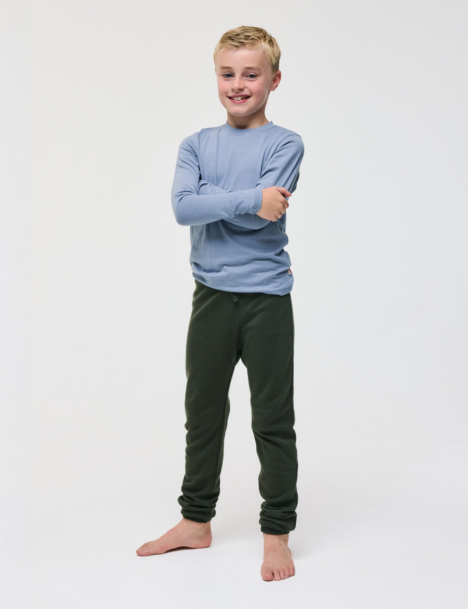 A smiling child stands barefoot against a white background, with arms crossed, wearing a long-sleeve light blue shirt and dark green Merino Kids AirWool sweatpants.