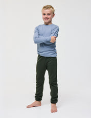 A smiling child stands barefoot against a white background, with arms crossed, wearing a long-sleeve light blue shirt and dark green Merino Kids AirWool sweatpants.