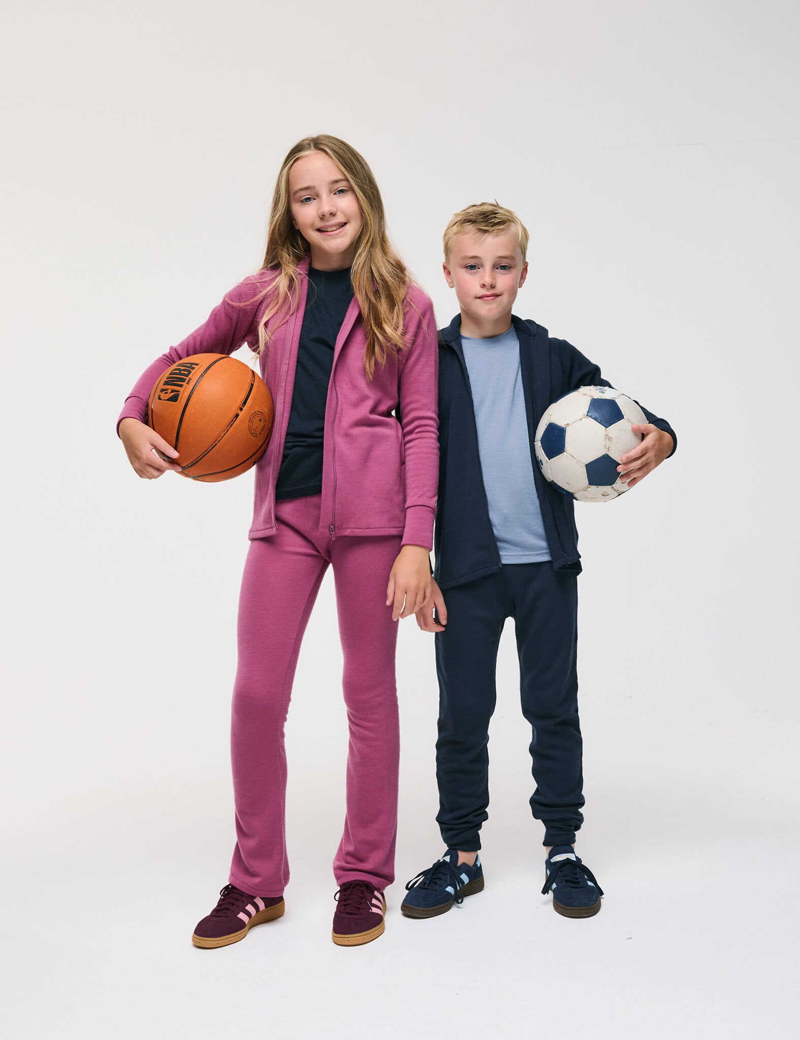 A girl in the Flarepants – Merino Kids AirWool tracksuit in soft pink holds a basketball, while a boy in the Flarepants – Merino Kids AirWool tracksuit in blue, made with comfortable brushed AirWool material, holds a soccer ball. Both are smiling and standing side by side against a plain white background.