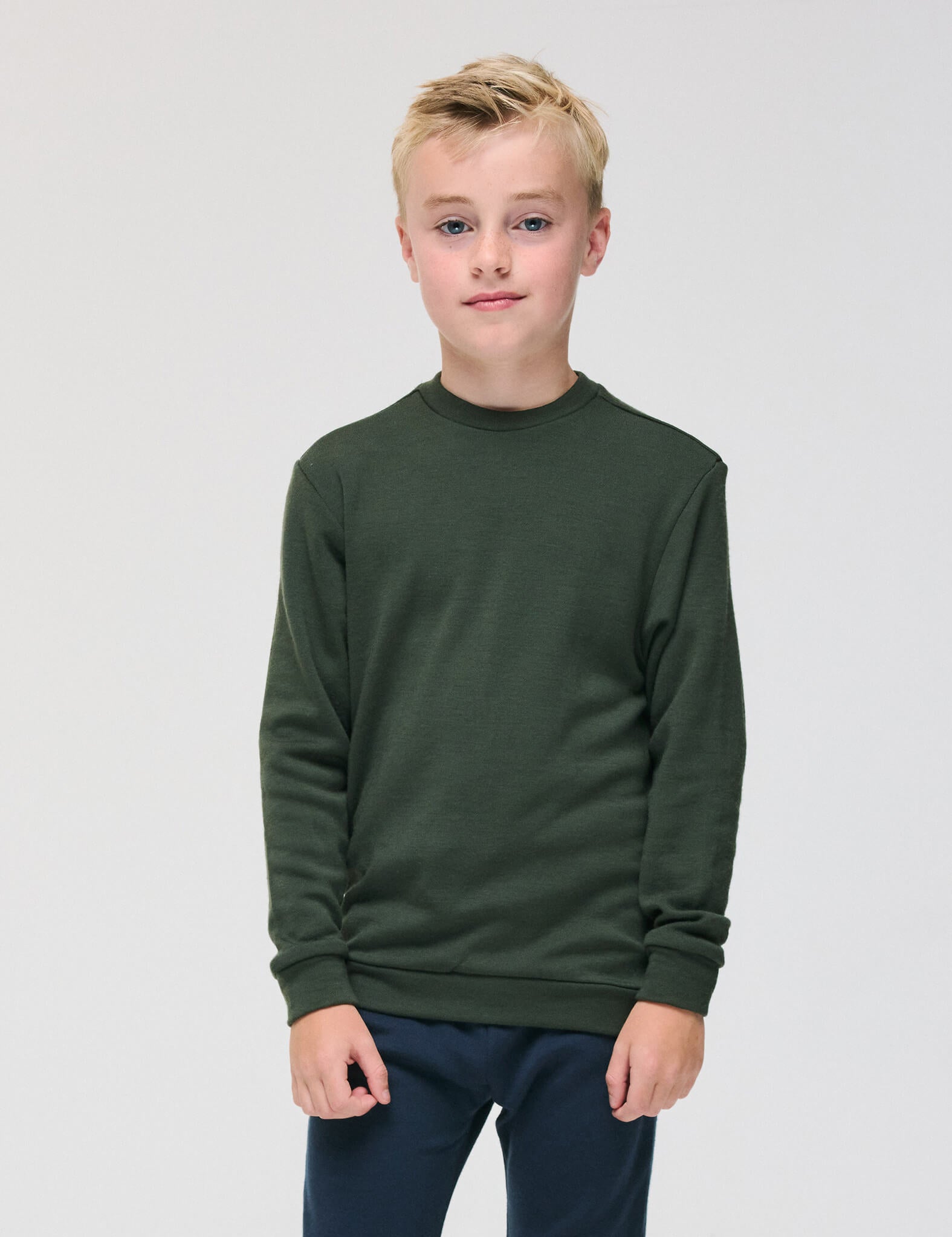 A young boy with short blond hair stands against a plain background, wearing the dark green Pullover – Merino Kids AirWool and navy pants. He is facing forward with a neutral expression.