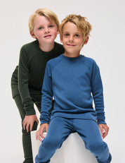 Two young children wear Pullover – Merino Kids AirWool sets, featuring long-sleeved tops and pants crafted from soft merino wool—one in green, one in blue—as they pose together against a plain white background. The child in blue is seated while the child in green stands behind.