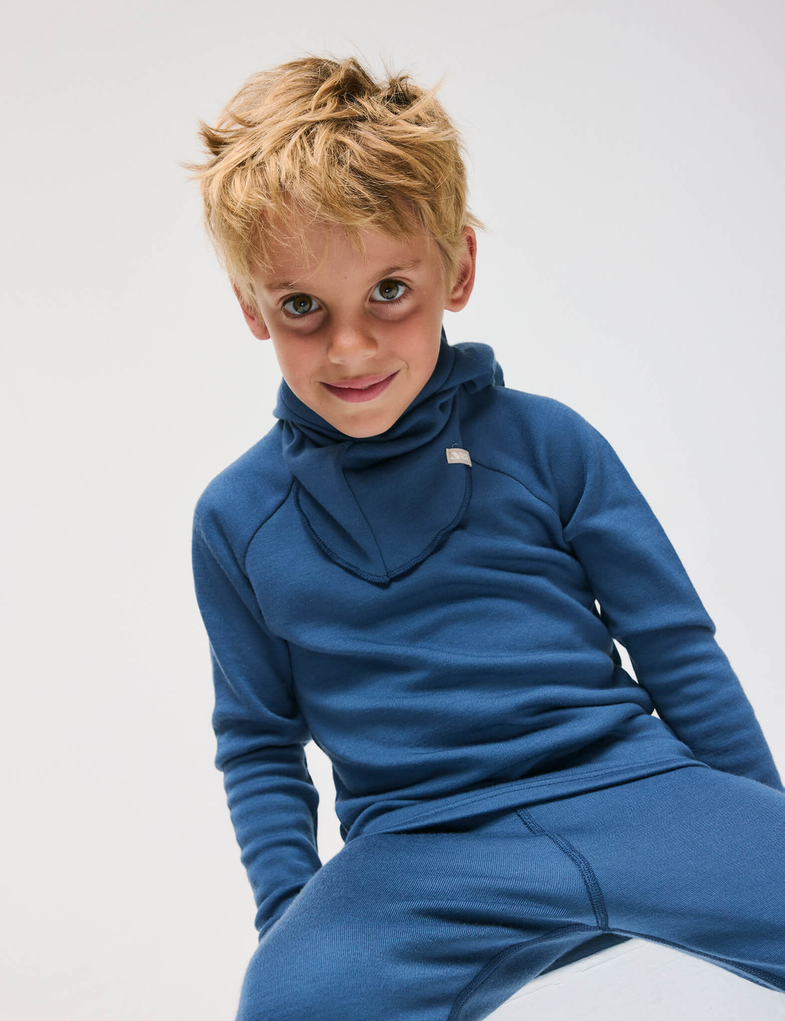 A young boy with tousled blond hair wears the Ensign Blue Pullover – Merino Kids AirWool sweater and matching pants, sitting and smiling at the camera against a plain light background.
