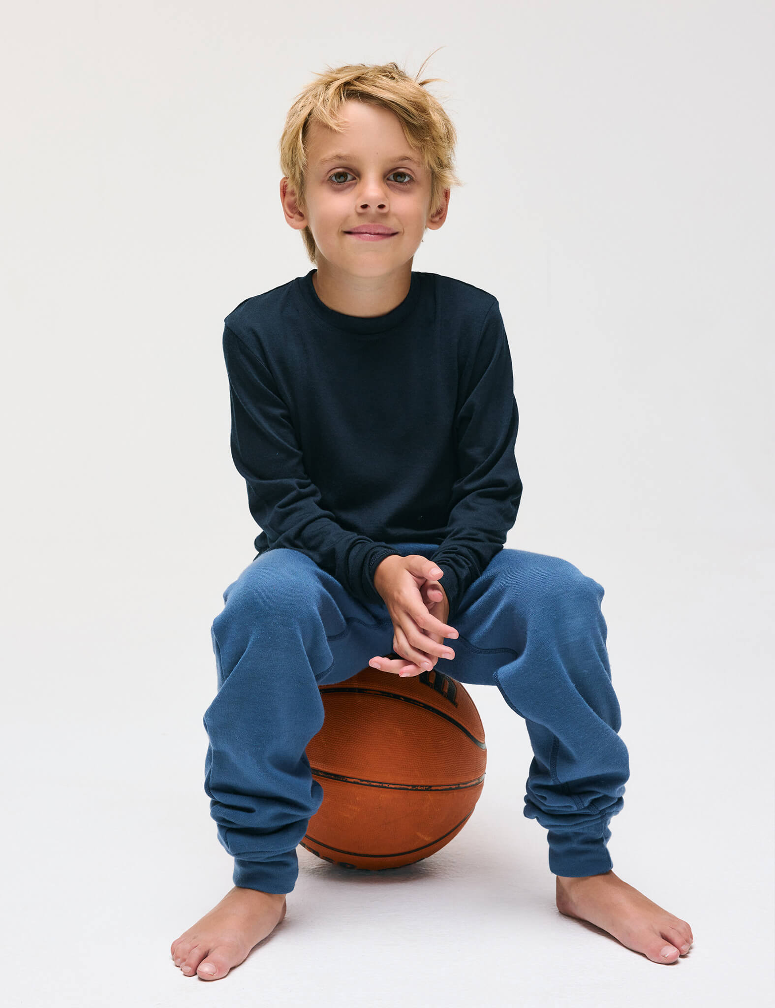 A young boy with blond hair sits barefoot on a basketball, smiling at the camera against a plain white background while wearing the Merino Kids AirWool Pullover in navy and blue sweatpants.