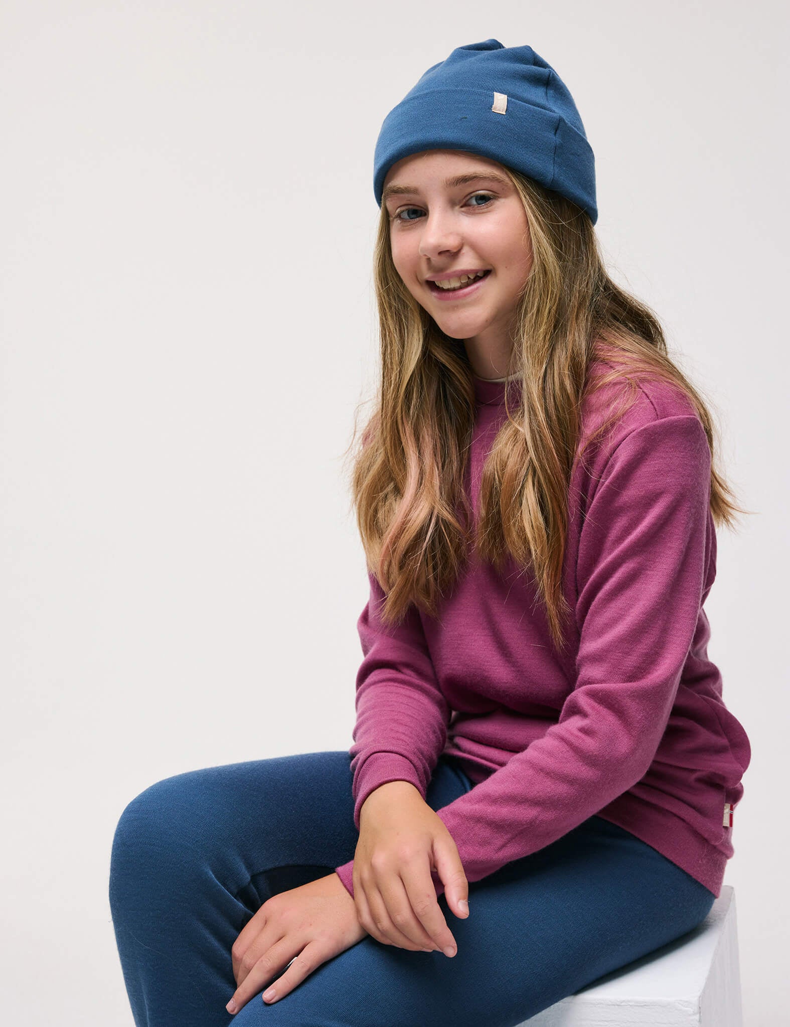 A girl with long hair sits on a chair, smiling. She wears a blue beanie, blue pants, and the Pullover – Merino Kids AirWool in Hawthorn Rose. The background is plain white.
