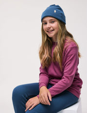 A girl with long hair sits on a chair, smiling. She wears a blue beanie, blue pants, and the Pullover – Merino Kids AirWool in Hawthorn Rose. The background is plain white.