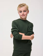 A young boy with short blond hair stands with his arms crossed, smiling slightly, against a plain light background. He is wearing the Pullover – Merino Kids AirWool in Forest Night.