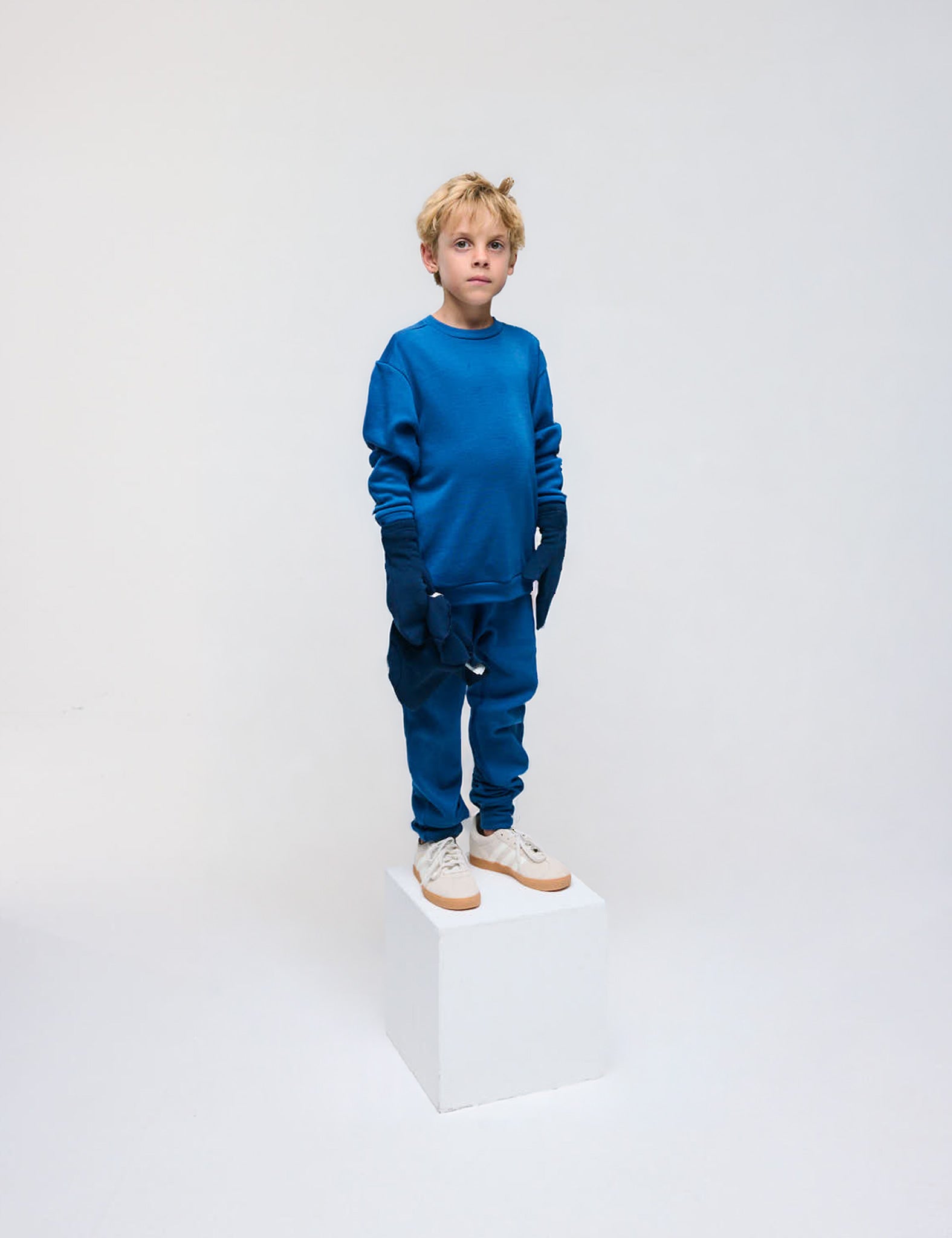 A young boy in a blue Pullover – Merino Kids AirWool sweater and matching pants stands on a white cube against a plain white background, wearing light-colored sneakers and dark gloves.