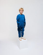 A young boy in a blue Pullover – Merino Kids AirWool sweater and matching pants stands on a white cube against a plain white background, wearing light-colored sneakers and dark gloves.