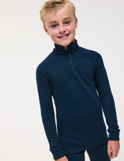 A smiling child with short blond hair wears the Half Zip – Merino Kids AirWool shirt in dark navy blue, paired with matching pants, standing against a plain light background.