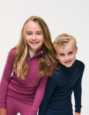 Two smiling children pose together against a plain white background. The girl on the left wears a long-sleeve pink outfit, while the boy on the right wears the Half Zip – Merino Kids AirWool, a navy zip-up polo shirt crafted from soft merino wool.