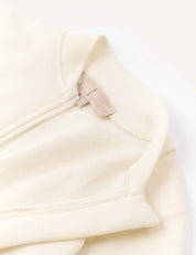 A close-up of the Onesie – Merino Baby AirWool showcases part of the collar, an exposed zipper, and a beige fabric tag with text, all set against a light background. The onesie features fold-over cuffs for added comfort.