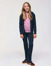 A young person stands smiling, wearing a pink shirt, navy hoodie, and matching navy pants with maroon sneakers, layered with the Merino Kids AirWool jacket against a plain white background.