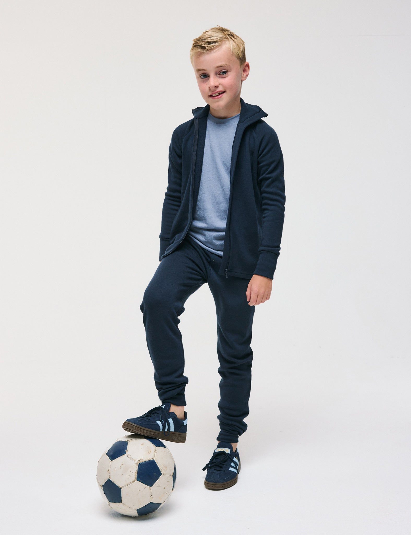 A young boy in a blue tracksuit and sneakers smiles at the camera as he stands with one foot on a black-and-white soccer ball against a plain white background, wearing the Merino Kids AirWool jacket.