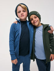 Two children, both wearing beanies and the Merino Kids AirWool zip-up jackets, stand close together with one arm around each other, smiling at the camera against a plain white background.