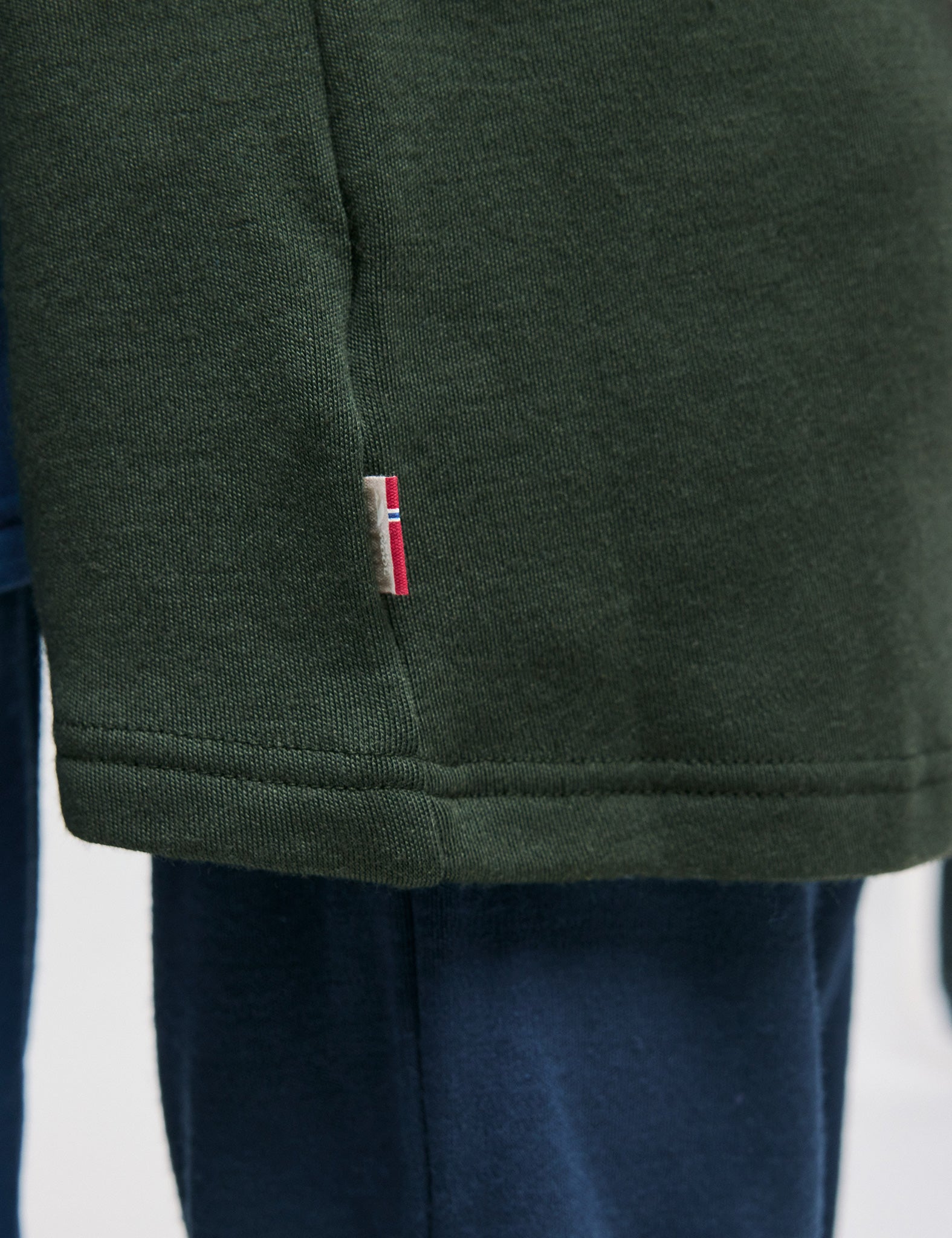 A close-up of the hem of a dark green shirt with a small red, white, and blue tag, styled with dark blue pants—perfectly complemented by the classic Merino Kids AirWool Jacket.