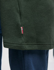 A close-up of the hem of a dark green shirt with a small red, white, and blue tag, styled with dark blue pants—perfectly complemented by the classic Merino Kids AirWool Jacket.