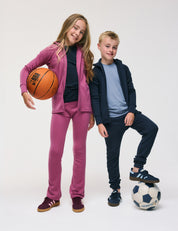 Two kids smile against a white background; the girl in pink holds a basketball while the boy in navy rests his foot on a soccer ball. Both are dressed in casual sportswear, with the boy wearing the Jacket – Merino Kids AirWool for extra comfort.